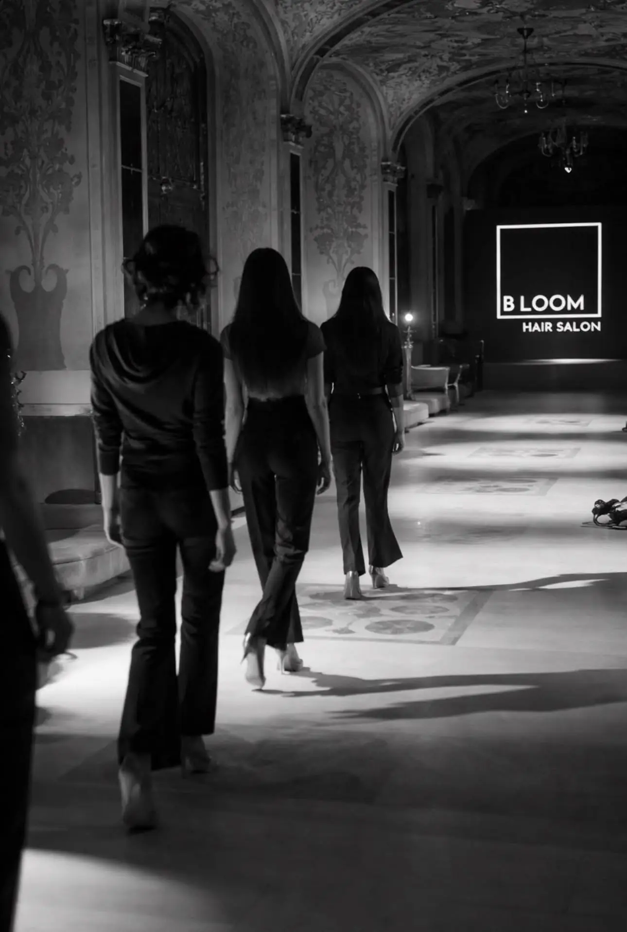 Modele pe podium într-o sală ornamentată, cu logoul BLOOM Hair Salon proiectat pe peretele din fund, fotografie alb-negru